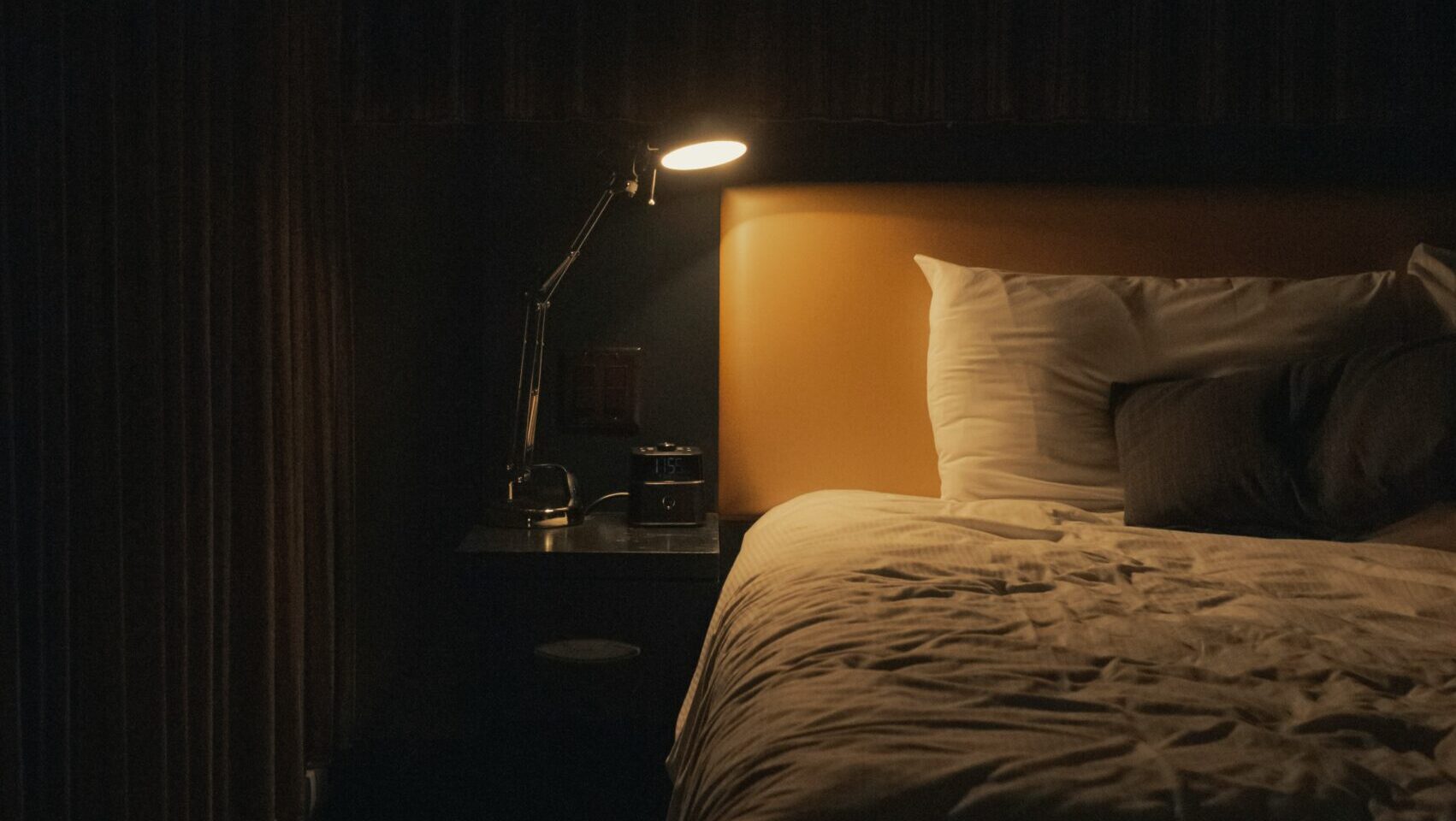 Uma cama de casal arrumada e organizada no centro de um quarto moderno e aconchegante à noite. O ambiente está imerso em uma penumbra suave, iluminado de forma indireta por fitas de LED de luz amarela quente posicionadas atrás da cabeceira e sob as prateleiras decorativas da parede lateral. Lençóis e travesseiros em tons cinza-claros e beges transmitem uma sensação extrema de conforto, ordem e relaxamento, ideal para um descanso profundo.