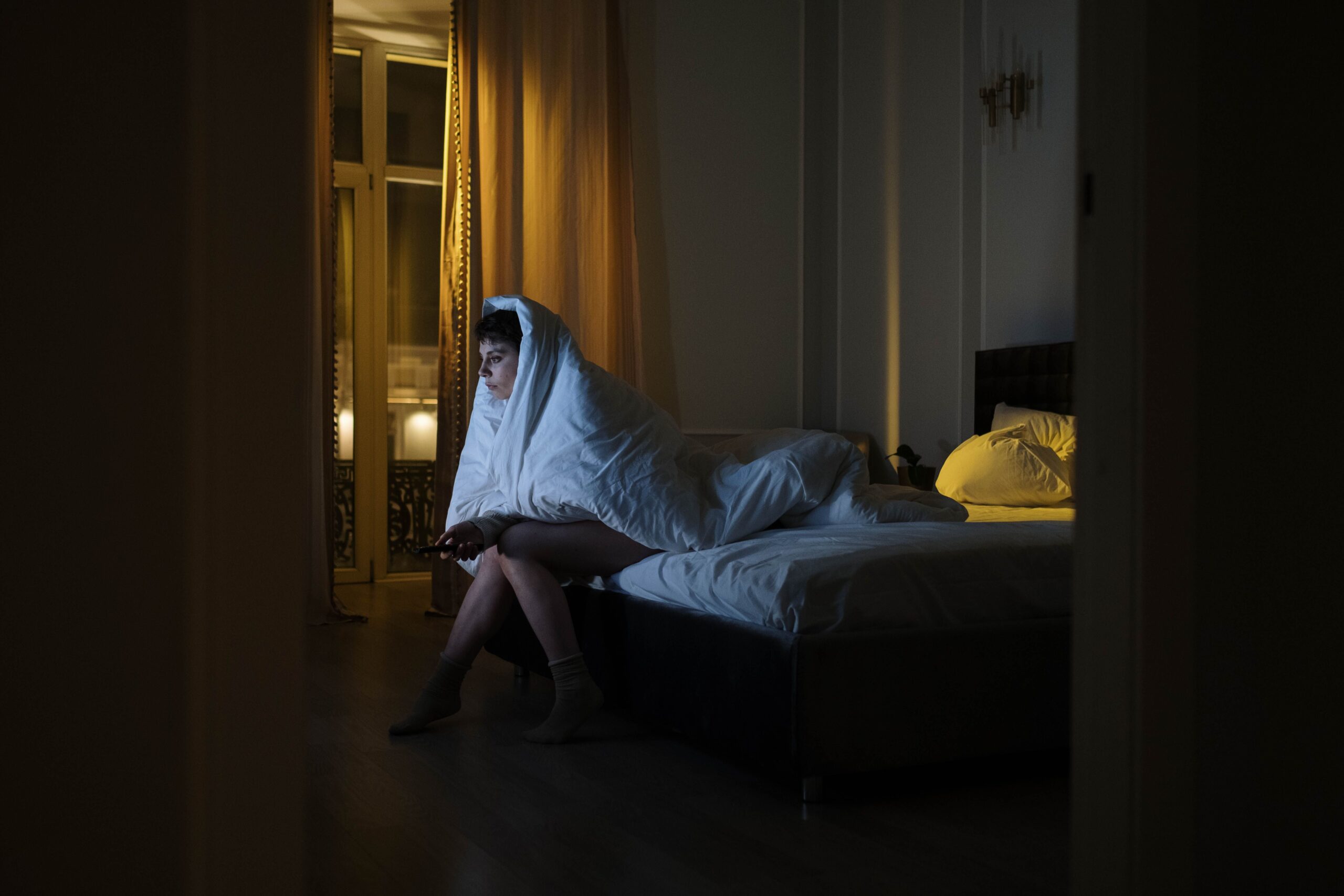 celular, café e televisão acesa representando hábitos que prejudicam o sono. Uma mulher jovem está sentada na beira de uma cama de casal em um quarto escuro à noite, enrolada em um edredom branco que cobre sua cabeça e corpo. Ela segura um controle remoto e olha fixamente para frente na penumbra. O ambiente é fracamente iluminado por uma luz amarela suave vinda de uma grande janela com cortinas ao fundo e de um abajur ao lado da cabeceira escura.